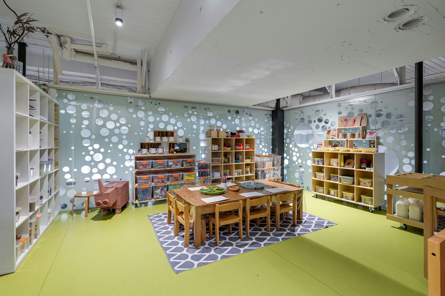Sala de arte infantil con mesas de madera y materiales de manualidades en estantes.